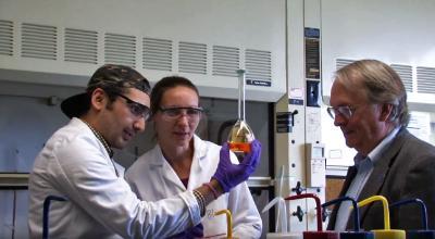 Binghamton University professor Stan Whittingham stands with 2 students who are examining a beaker filled with liquid.
